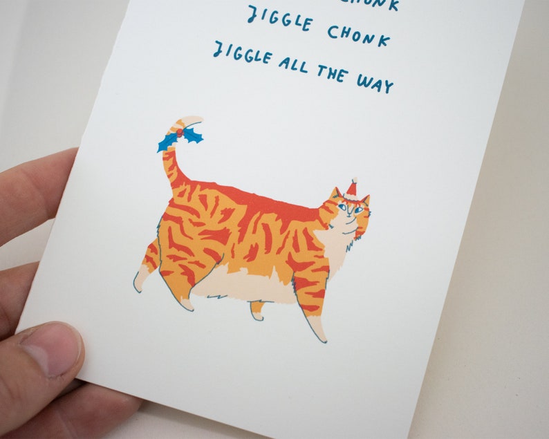 May include: A greeting card featuring a cartoon orange tabby cat wearing a red Santa hat with a blue trim. The cat has a white chest and belly. The text on the card reads "Jiggle Chonk Jiggle All The Way".