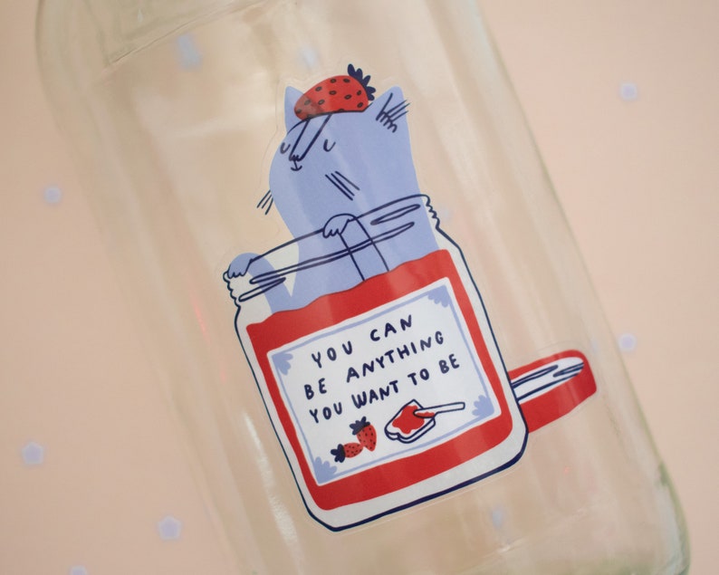 May include: A glass jar with a red and white sticker featuring a cartoon cat wearing a strawberry on its head. The sticker says "You can be anything you want to be".