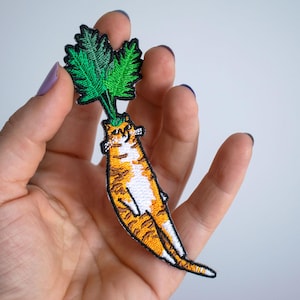May include: Embroidered patch with a quirky design of a cat shaped like a carrot. The cat is orange and white with green leaves at the top. The patch is held in a hand against a white background.