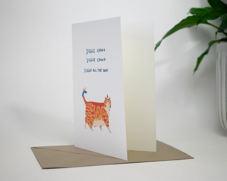 May include: A greeting card with a cartoon orange cat wearing a blue party hat. The text on the card reads "Jiggle Chonk Jiggle Chonk Jiggle All The Way".