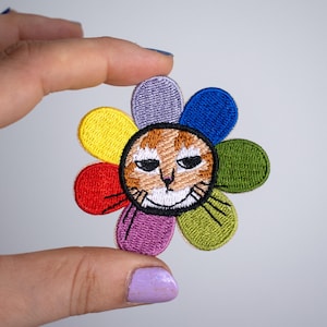 Può includere: Toppa ricamata con la faccia di un gatto al centro di un disegno floreale. I petali del fiore sono di vari colori: rosso, giallo, viola, blu e verde. Il gatto ha il pelo arancione e bianco e un'espressione leggermente scontrosa.