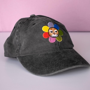 Peut inclure: Casquette de baseball gris anthracite avec une visière incurvée. Le chapeau présente un motif brodé d'un visage de chat au centre d'une fleur colorée. Les pétales de la fleur sont dans les tons de jaune, rouge, rose, violet, bleu et vert.