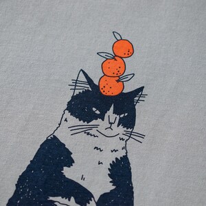 May include: A light gray t-shirt featuring a whimsical illustration of a black and white cat with three orange tangerines stacked on its head. The cat has a slightly grumpy expression.