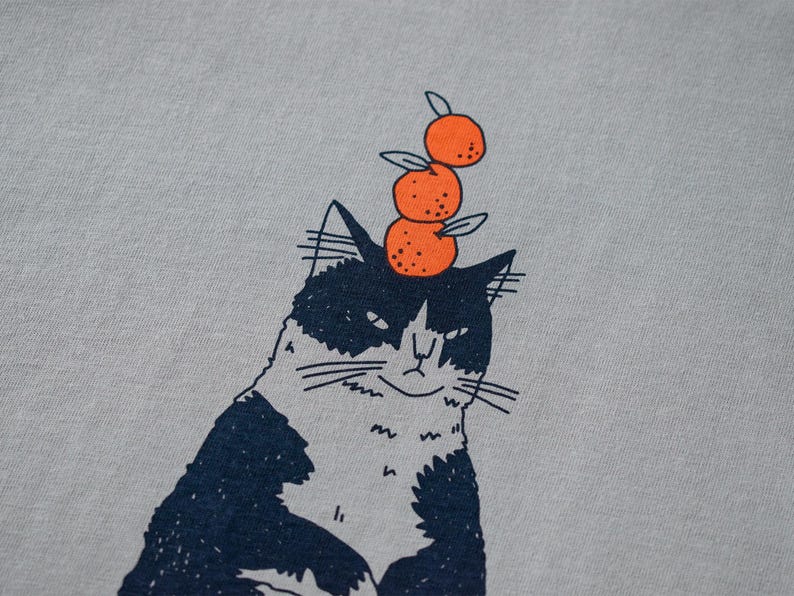May include: A light gray t-shirt featuring a whimsical illustration of a black and white cat with three orange tangerines stacked on its head. The cat has a slightly grumpy expression.