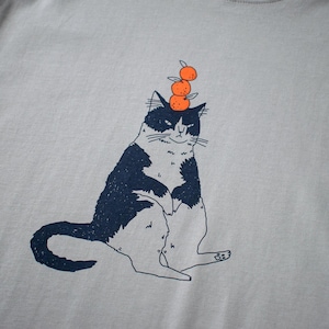 May include: Light gray t-shirt featuring a whimsical illustration of a black and white cat balancing three orange fruits on its head. The cat is sitting upright with a slightly grumpy expression. The design is printed in navy blue and orange.