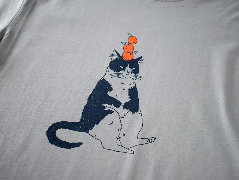 May include: Light gray t-shirt featuring a whimsical illustration of a black and white cat balancing three orange fruits on its head. The cat is sitting upright with a slightly grumpy expression. The design is printed in navy blue and orange.