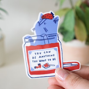 May include: A blue cat wearing a strawberry hat is inside a jar of red jam. The jar has a white label with the text "You can be anything you want to be".