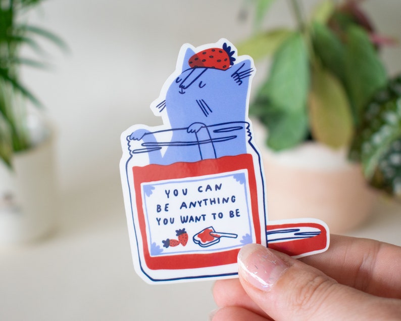 May include: A blue cat wearing a strawberry hat is inside a jar of red jam. The jar has a white label with the text "You can be anything you want to be".