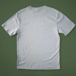 May include: A light gray, short-sleeved t-shirt, viewed from the back, lies on a green surface. The shirt has a crew neck and a simple, classic design, suitable for casual wear.