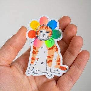 Mürrische Blumenkatze | Holo-Vinyl Sticker einer leicht orange getigerten Katze, die ein regenbogenfarbenes Blütenblatt-Kostüm um den Kopf trägt