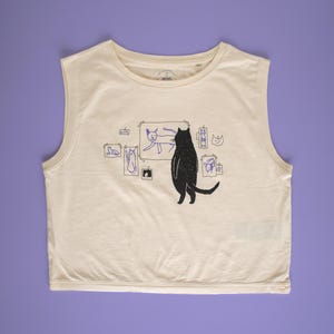 Op de afbeelding: Crèmekleurige mouwloze crop top met een zwarte kattenillustratie. De kat kijkt naar een ingelijste tekening van een kat. Extra blauwe kattentekeningen worden rond de hoofdafbeelding weergegeven. De top is gemaakt van een zachte, lichte stof.