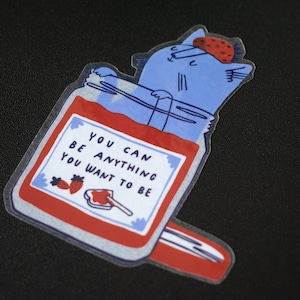 May include: A blue cat with a strawberry on its head is inside a red jar with a white label that says "You can be anything you want to be".