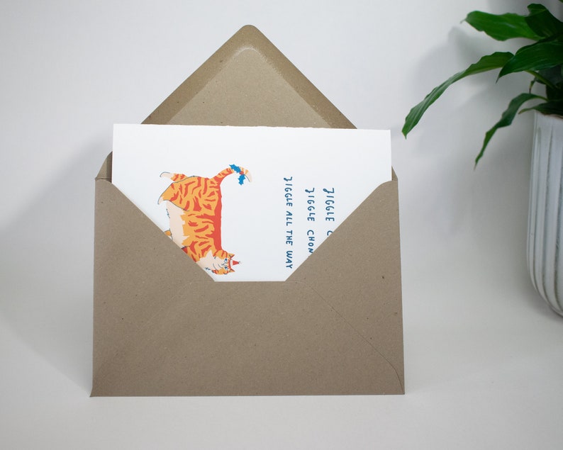 May include: A greeting card with a cartoon illustration of an orange and white cat with blue eyes. The cat is walking with its tail up in the air. The text on the card reads "Jiggle Jiggle Chonky Jiggle All The Way".