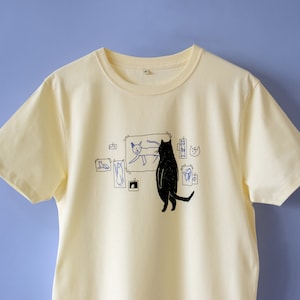 Kat galerij t-shirt | Handzeefdruk op ecru/lichtgeel T-shirt van biologisch katoen met zwart-blauwe illustratie van een kat die kattenkunst beoordeelt