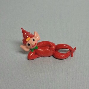 May include: A small, red figurine of a whimsical elf or gnome. The figure is lying down, with a pointed hat covered in white polka dots. It has a green bow tie and a smiling face. The figurine is made of a smooth material.