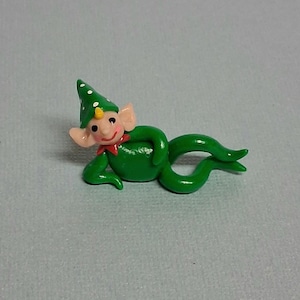 May include: A whimsical green elf figurine with a pointed hat adorned with white polka dots. The elf has a smiling face, pink ears, and a red collar. The figurine is lying down with its legs curled.
