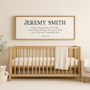 May include: A white framed print with the name "JEREMY SMITH" and a bible verse from Jeremiah 1:5. The print is hanging on a white wall above a wooden crib.