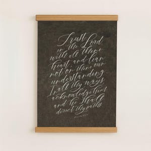 May include: A framed wall art print with a dark grey background and white calligraphy script. The text reads "Trust Lord in the thine with all thine heart and lean not on thine understanding." The frame is made of light brown wood.