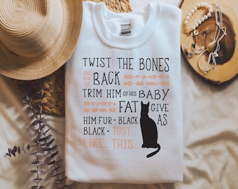 May include: White sweatshirt with black text that reads "Twist the Bones and Bend the Back, Trim him of his Baby Fat, Give him fur: Black as Black...Just like this..." with a black cat illustration.