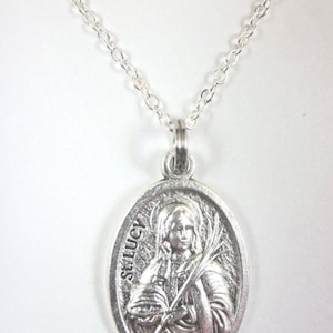 May include: Silver-coloured oval pendant necklace with a detailed relief of a saint, inscribed with "St. Lucy". The pendant hangs from a delicate chain. The saint is shown holding a palm branch and a globe.