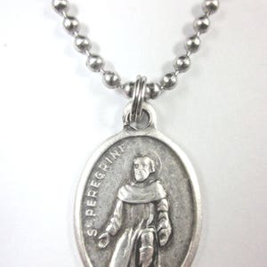 May include: Silver-coloured pendant necklace featuring an oval pendant with an image of a saint. The pendant is suspended from a ball chain necklace. The pendant has the words "S. PEREGRINE" around the image.