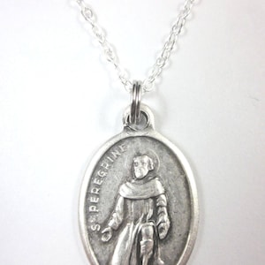 May include: A silver pendant necklace with an oval pendant depicting St. Peregrine. The pendant hangs from a fine silver chain. The saint is shown kneeling, with the inscription "S. PEREGRINE" around the edge.
