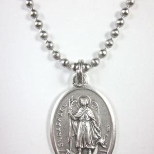 Op de afbeelding: Een zilveren ketting met een zilveren ovale hanger met een afbeelding van Sint Rafaël. De hanger heeft de woorden "St. Raphael" erop gegraveerd.