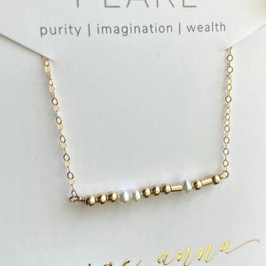 Op de afbeelding: Een gouden ketting met een staaf van gouden kralen en parels. De ketting ligt op een witte kaart met de tekst "PEARL puurheid | verbeelding | rijkdom" en "paige anna jewelry".