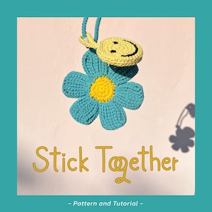 Puede incluir: Un colgante de crochet con forma de flor azul con centro amarillo y una carita sonriente amarilla. El texto "Stick Together" está en dorado, con "- Pattern and Tutorial -" debajo.