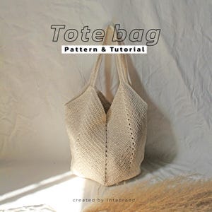 Puede incluir: Una bolsa de mano de crochet beige con una abertura en forma de V. La bolsa está colgada en una pared blanca con el texto "Tote bag Pattern & Tutorial" encima. El texto "created by intabrand" está debajo de la bolsa.