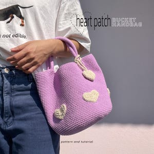 May include: A lavender crochet bucket handbag featuring cream-colored heart-shaped patches and a matching heart charm attached to the handle. The text heart patch BUCKET HANDBAG and pattern and tutorial are visible on the image.
