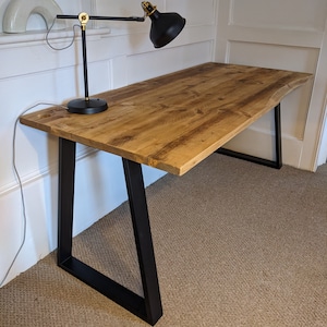 Op de afbeelding: Een rustiek houten bureau met een live edge blad en zwarte metalen poten. Het bureau heeft een natuurlijke houten afwerking en wordt ondersteund door twee A-vormige poten.