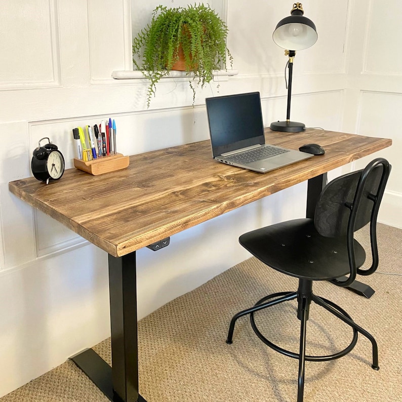 May include: A black standing desk with a wooden top and black metal legs. A black office chair with a black metal base is positioned in front of the desk. A laptop computer, a lamp, and a small alarm clock are on the desk.