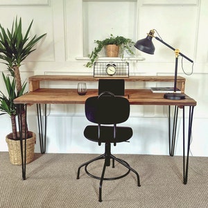 May include: A wooden desk with a black metal frame and hairpin legs. The desk has a shelf above the work surface. A black office chair with a black metal base is positioned in front of the desk. A black lamp with a gold base is on the desk. A potted plant is on the shelf.