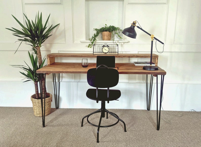 May include: A wooden desk with a black metal frame and hairpin legs. The desk has a shelf above the work surface. A black office chair with a black metal base is positioned in front of the desk. A black lamp with a gold base is on the desk. A potted plant is on the shelf.