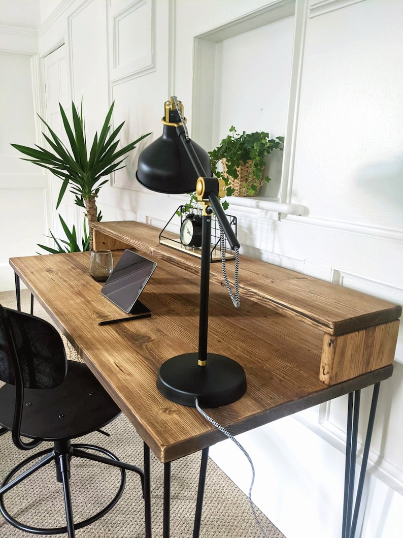 May include: A rustic wooden desk with black hairpin legs and a black desk lamp with a gold base. The desk has a shelf underneath the top surface. A tablet computer and a pen are on the desk.