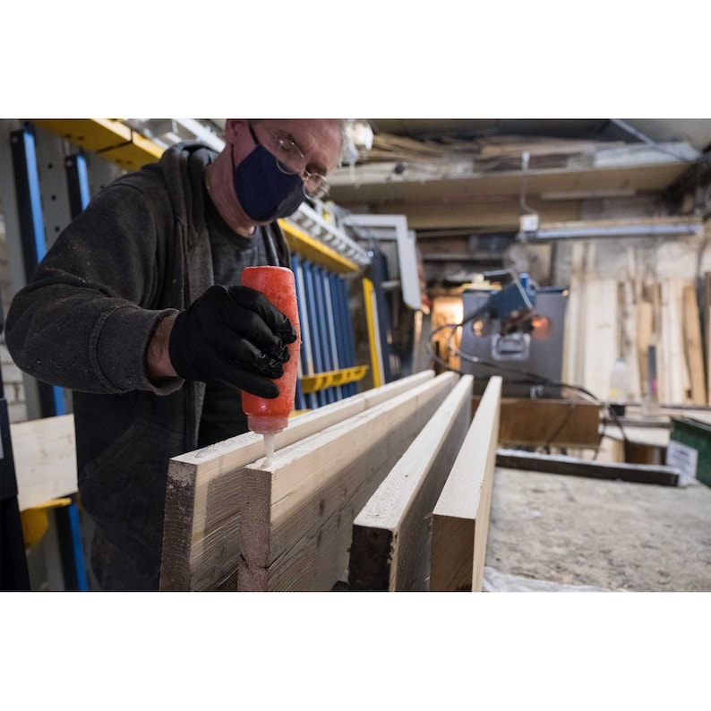 May include: A person wearing a black face mask and black gloves applies glue to a long piece of wood.