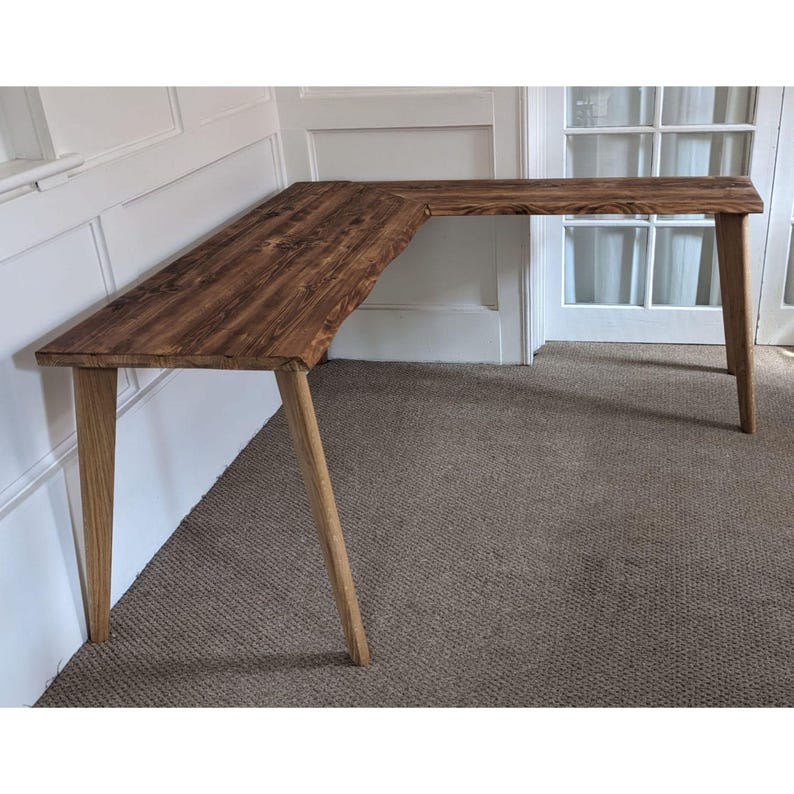 May include: A corner desk made of reclaimed wood with a natural brown finish. The desk has a simple design with four legs and a rectangular top. The desk is sitting on a carpeted floor.