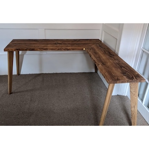 May include: A corner desk made of reclaimed wood with a dark brown stain. The desk has a simple design with angled legs and a wide, rectangular top.