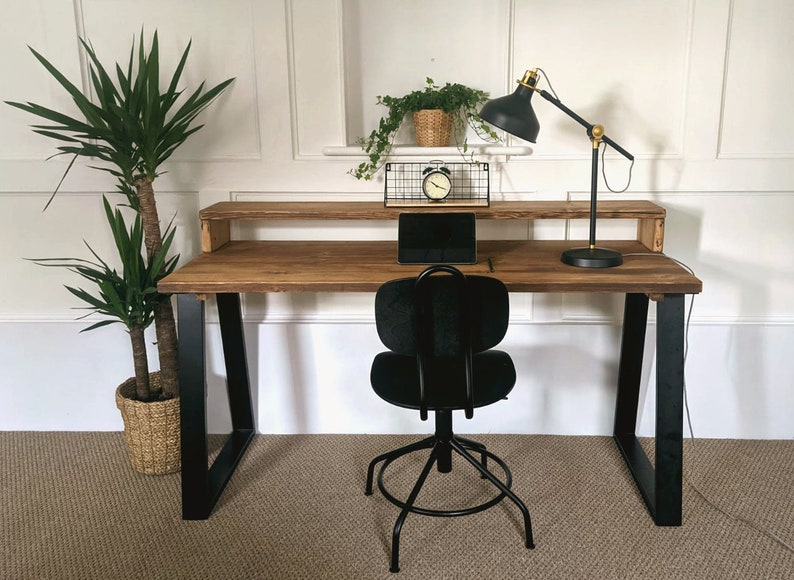 May include: A wooden desk with black metal legs and a black office chair. The desk has a shelf above the desktop. There is a lamp on the desk and a potted plant on the shelf.
