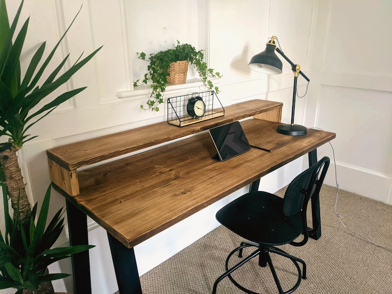 May include: A rustic wooden desk with black metal legs and a shelf. A black lamp with a gold accent sits on the desk, along with a tablet and a black alarm clock.