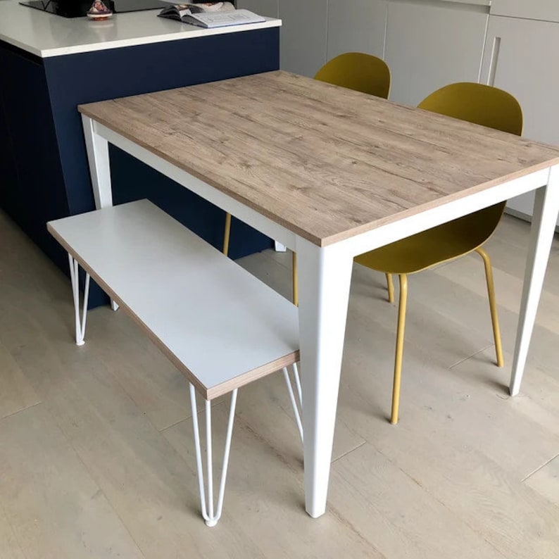 Puede incluir: Una mesa de comedor de madera blanca con una tapa de grano de madera y patas blancas. La mesa est&aacute; puesta con un banco blanco y dos sillas de color amarillo mostaza con patas doradas.
