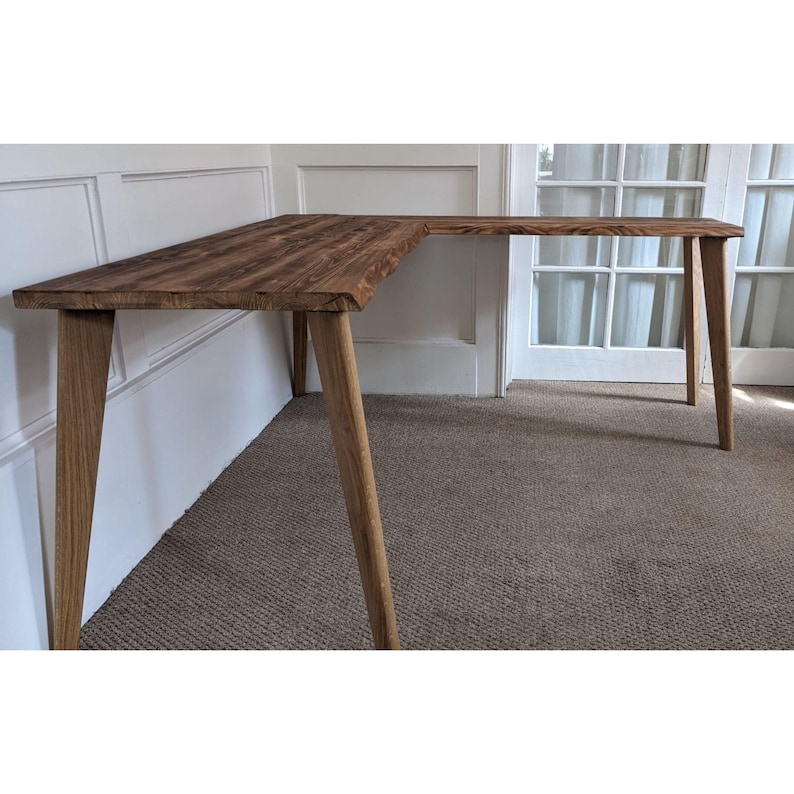May include: A wooden L-shaped desk with a natural wood finish and four legs. The desk is made of reclaimed wood and has a rustic look.