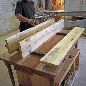 May include: Two long, light brown wooden planks are being measured and prepared for woodworking. The planks are resting on a workbench with a metal clamp holding them in place.