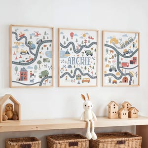 Op de afbeelding: Drie ingelijste kunstprints met een wegenkaartontwerp met voertuigen, gebouwen en bomen. De middelste print heeft de naam "Archie" in blauwe letters. De prints zitten in licht houten frames.