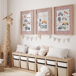 May include: Three framed wall prints featuring illustrations of construction vehicles, a police car, and a bicycle, with the name ARCHIE displayed on the center print. The prints have a red and white striped border and are hung above a wooden toy storage unit.