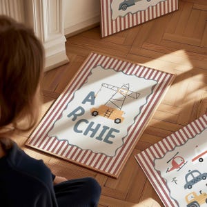 May include: A rectangular framed print featuring a white center with a scalloped edge, displaying the name ARCHIE in dark blue letters alongside a yellow construction crane illustration. The border has vertical red and white stripes.