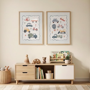 May include: Two framed wall art prints featuring illustrations of various vehicles including a helicopter, ambulance, tractor, police car, and construction equipment, displayed in light wood frames above a wooden storage unit with toys and books.