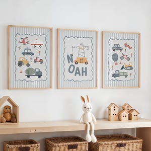 May include: Three framed wall art prints featuring illustrations of construction vehicles, cars, and a crane with the name NOAH in light wood frames. The prints have a gray and white striped background with a scalloped border around the graphics.