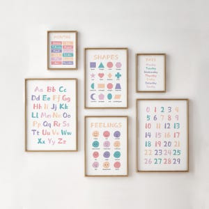 May include: A set of six framed educational prints with gold frames. The prints display the alphabet, shapes, months, days of the week, feelings, and numbers 0-29. The prints feature pastel colors and are arranged on a white wall.